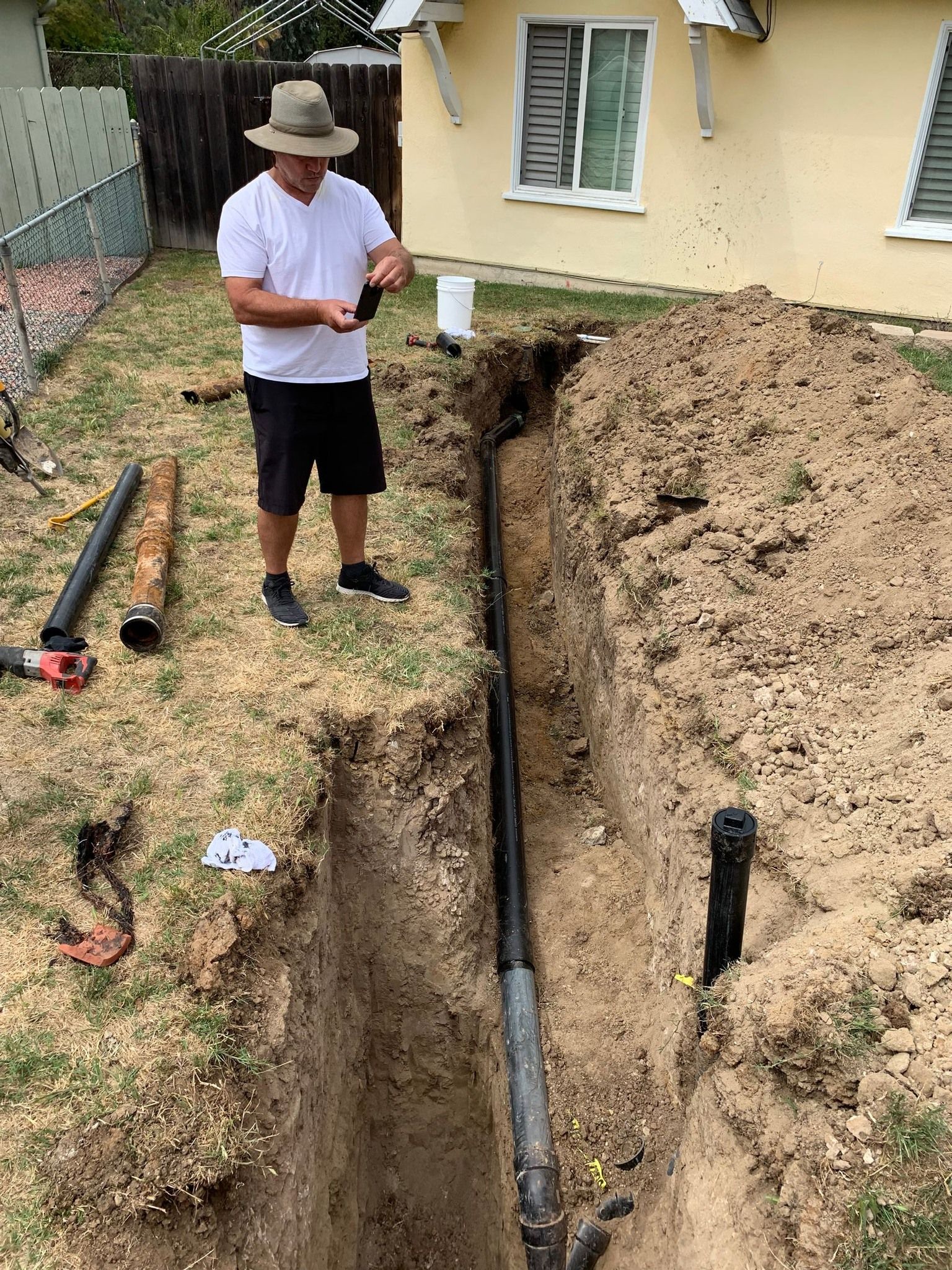 Man in hat by trench, working on black pipe in backyard.