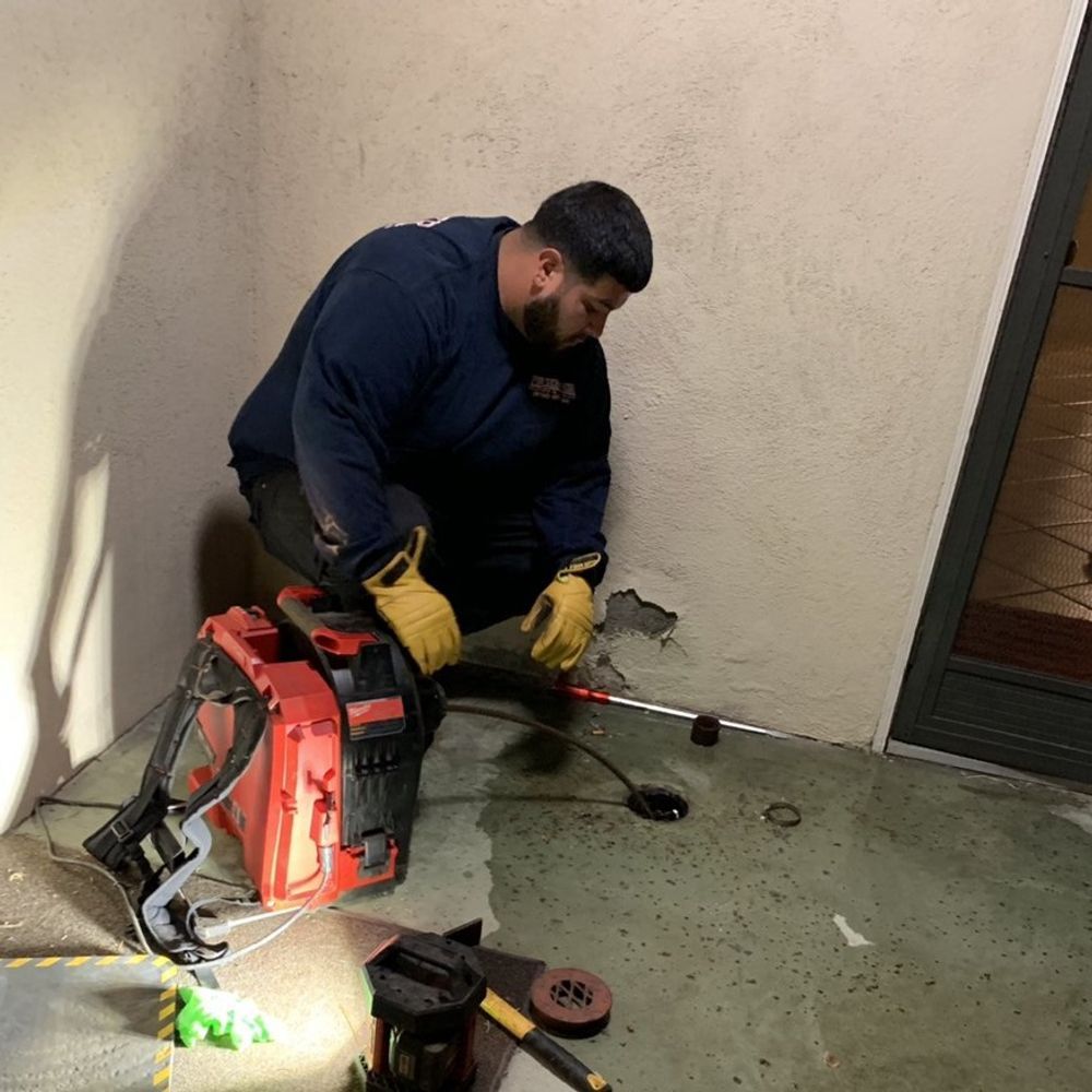 A person in work attire crouches next to a drain, using a tool. Setting is a corner of a building.