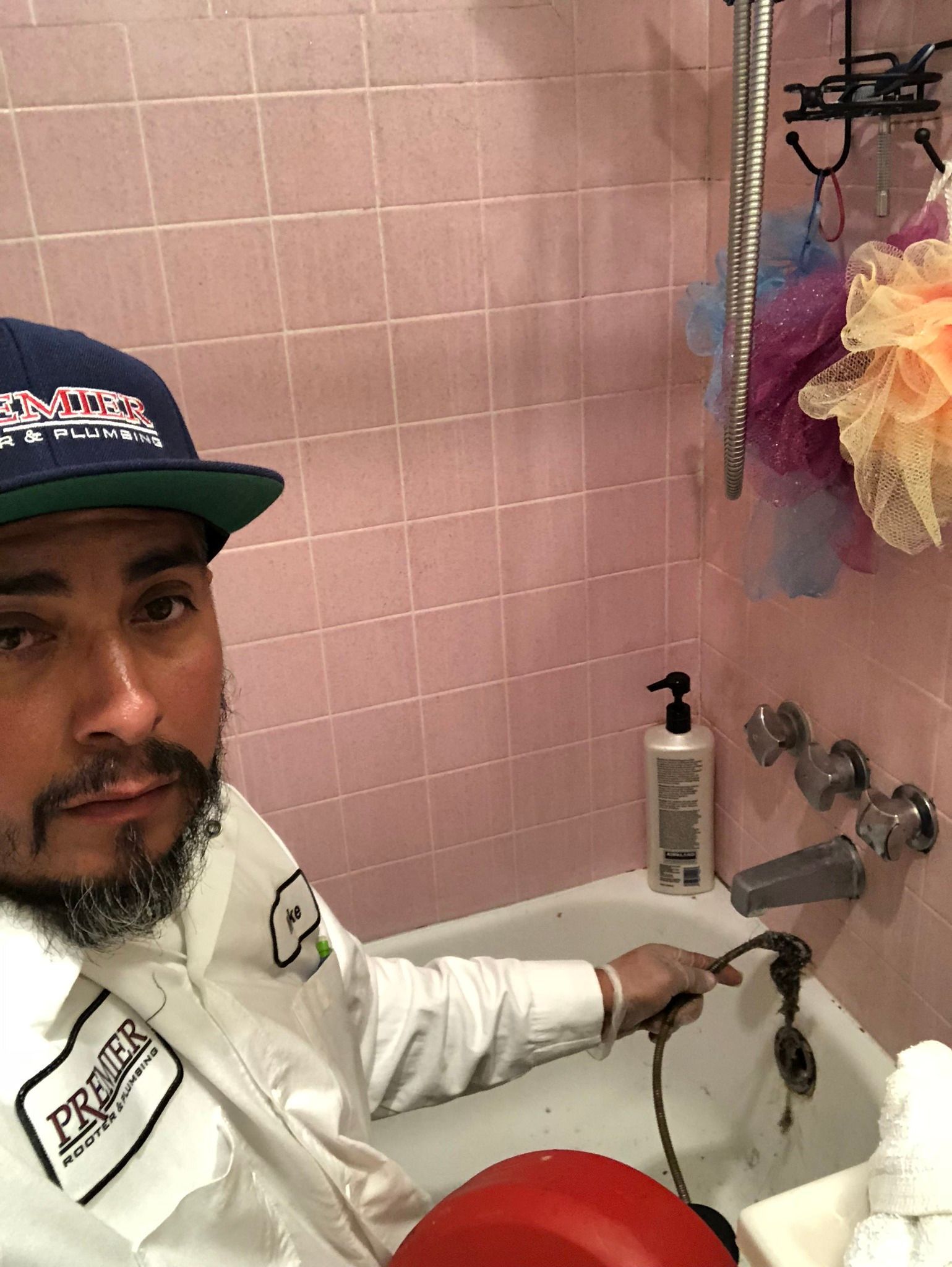 Plumber in bathtub inspecting faucet, pink tiled walls. He wears a cap and uniform.