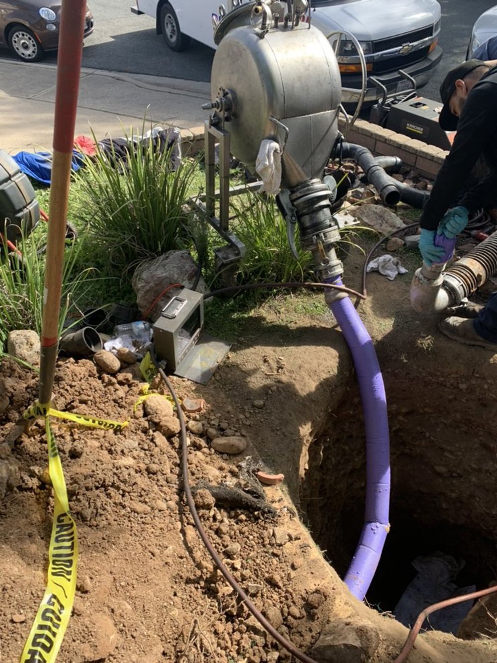A worker near an excavation with a large silver industrial machine and purple hose. Yellow caution tape surrounds the hole.