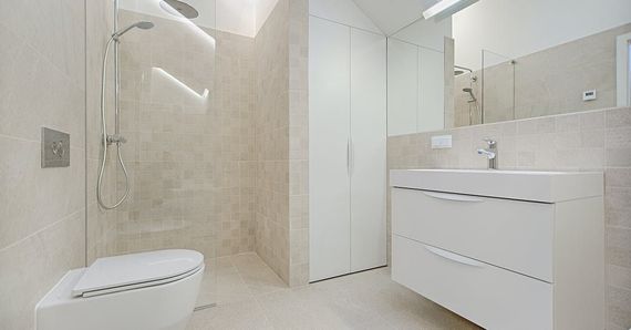 Modern bathroom with white cabinetry, glass shower, toilet, and sink. Beige tile walls and flooring.