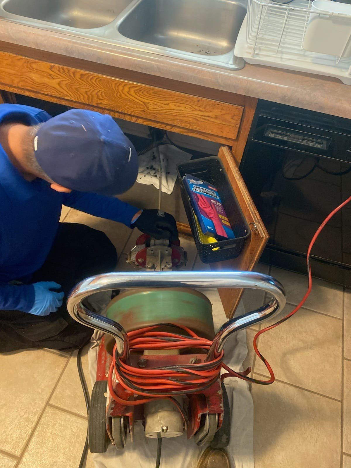 Plumber using a drain snake under a kitchen sink. Red machine with coiled cord. Person wears blue hat and gloves.