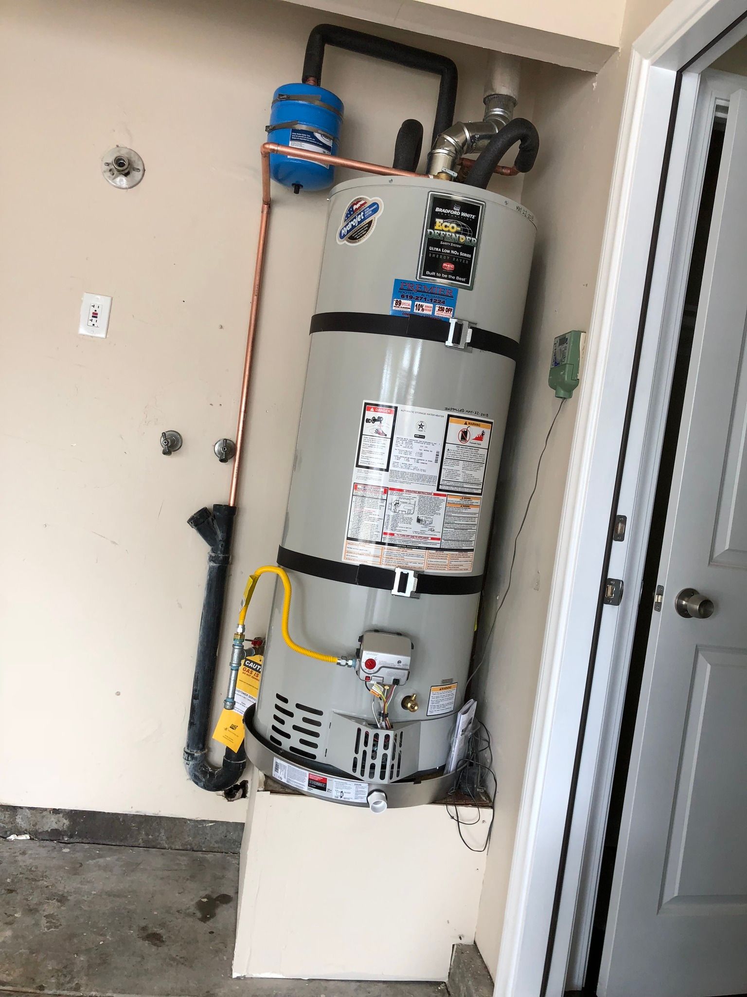 A water heater in a garage, next to a door. It's gray, with black straps. A blue tank is above it.