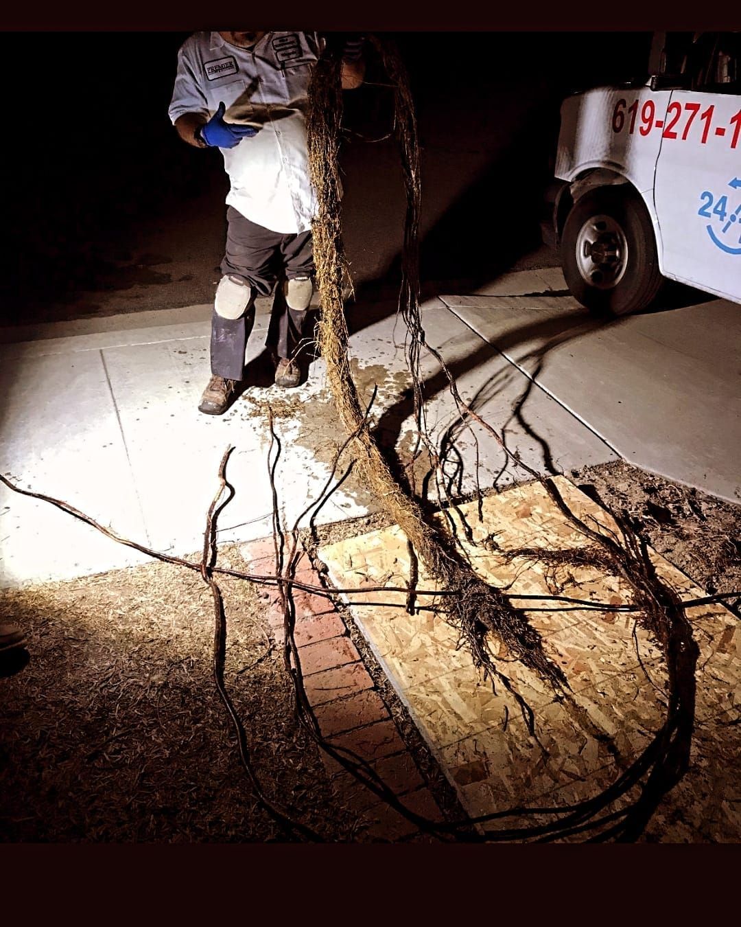 Person holding tree roots out of a hole in a sidewalk, lit by a flashlight, near a service truck.