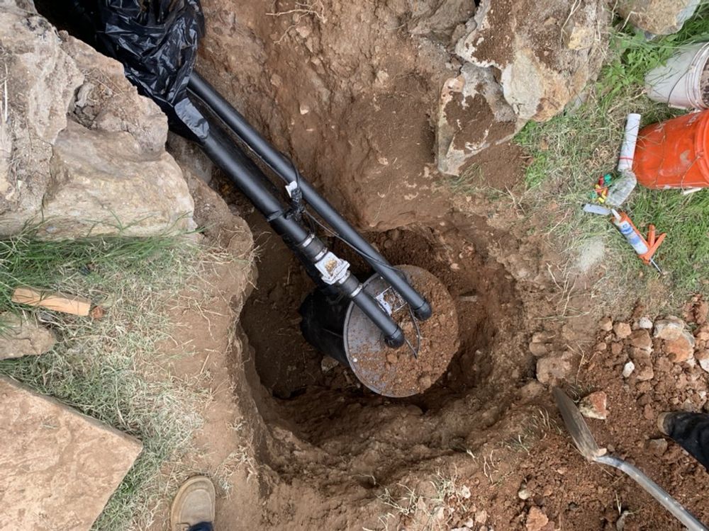 Hole in the ground with installed black pipe and round metal object. Tools and debris surround the hole.