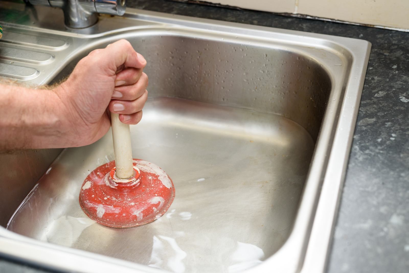 Person plunges a partially filled stainless steel sink with a red and white plunger.