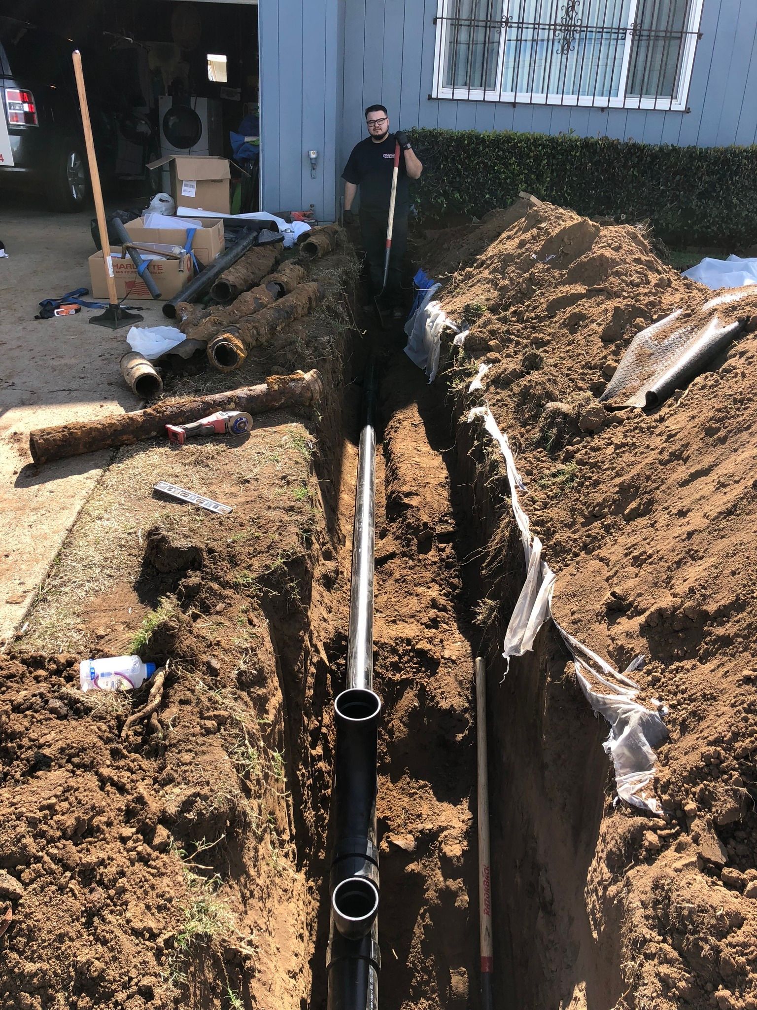 A worker in a trench with black pipes, preparing for plumbing work near a house.