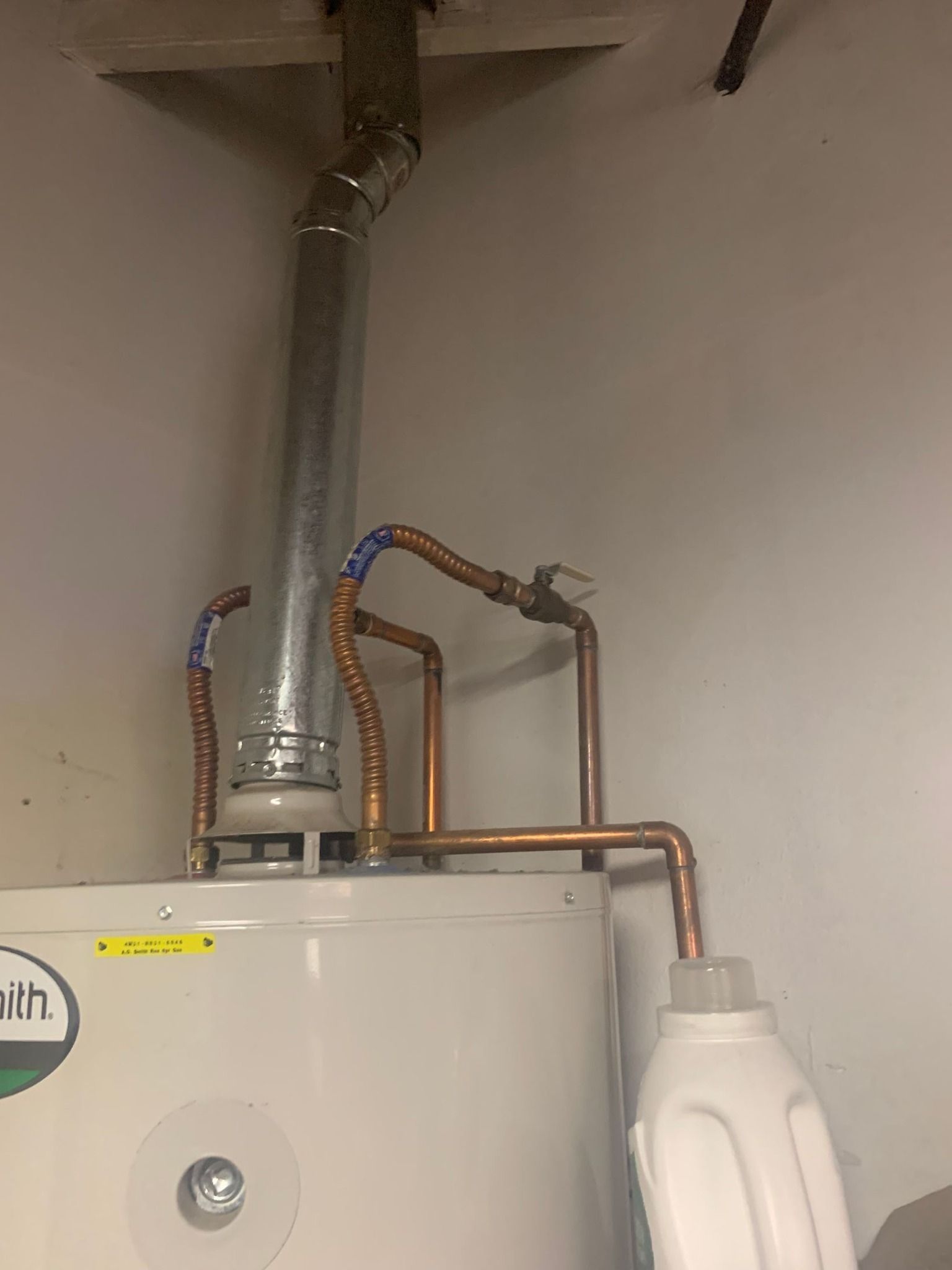 Water heater with copper pipes, exhaust vent, and a white plastic container attached.