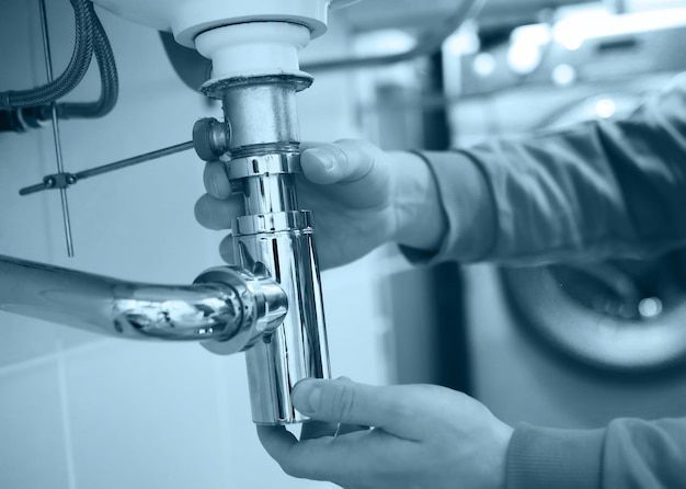 Person's hands assembling chrome plumbing under a sink. White walls, washing machine in the background.