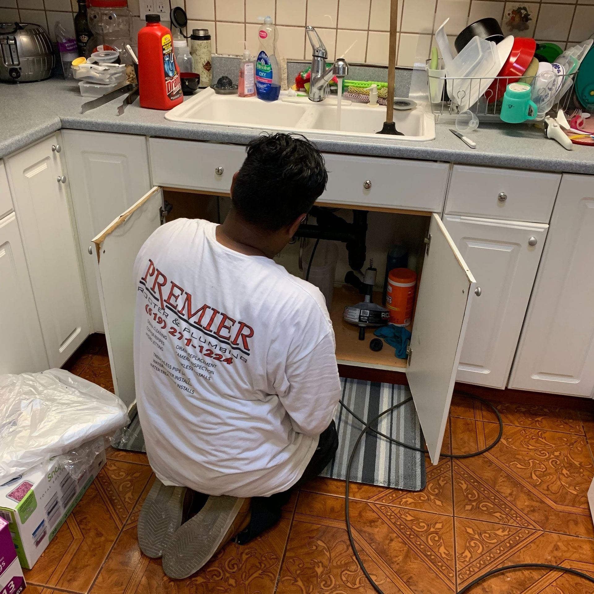 Person kneels to work under a kitchen sink. White cabinets, cluttered countertop, and tile floor are visible.