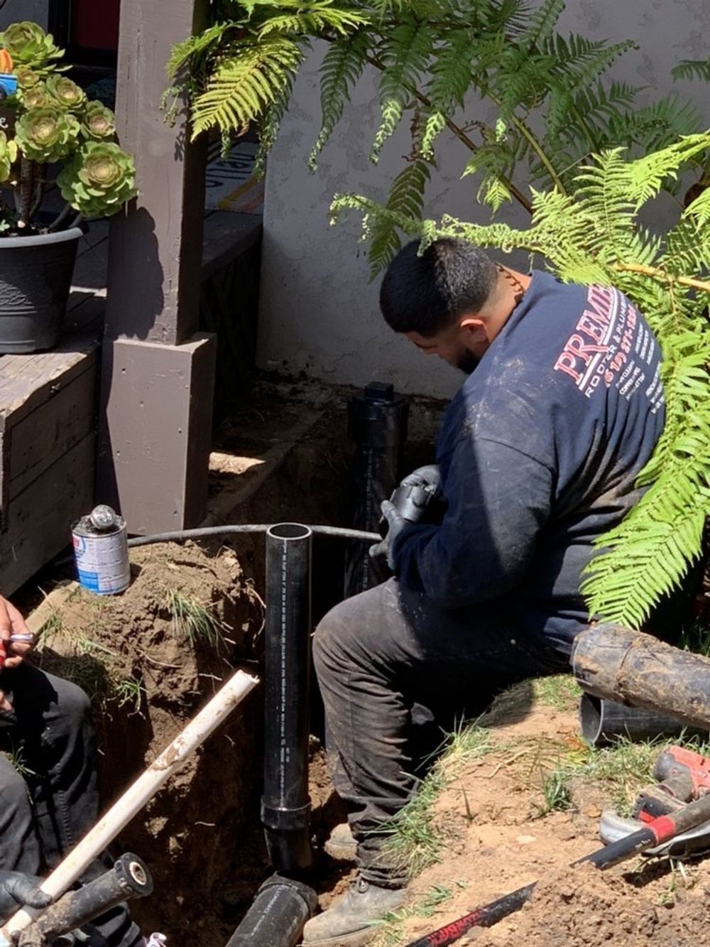 Person working under a kitchen sink. Dark cabinets, stainless steel sink. Tile floor, tools visible.