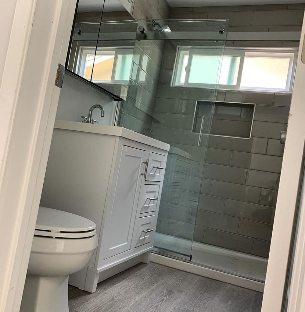 Bathroom with white vanity, toilet, glass shower, gray tile, and a window.
