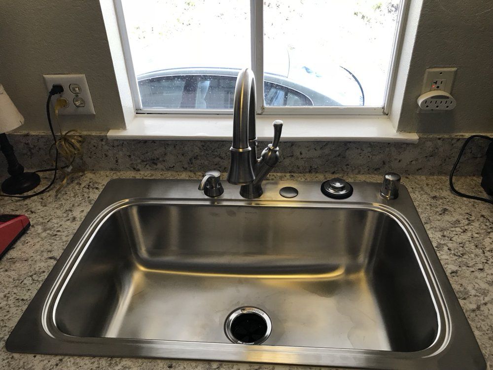 Stainless steel kitchen sink, faucet, soap dispenser, and window. Granite countertop with outlets nearby.