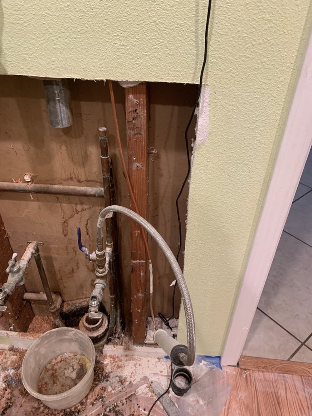 Plumbing exposed in a wall opening, including pipes, valves, and a bucket. Light green wall and wooden studs visible.