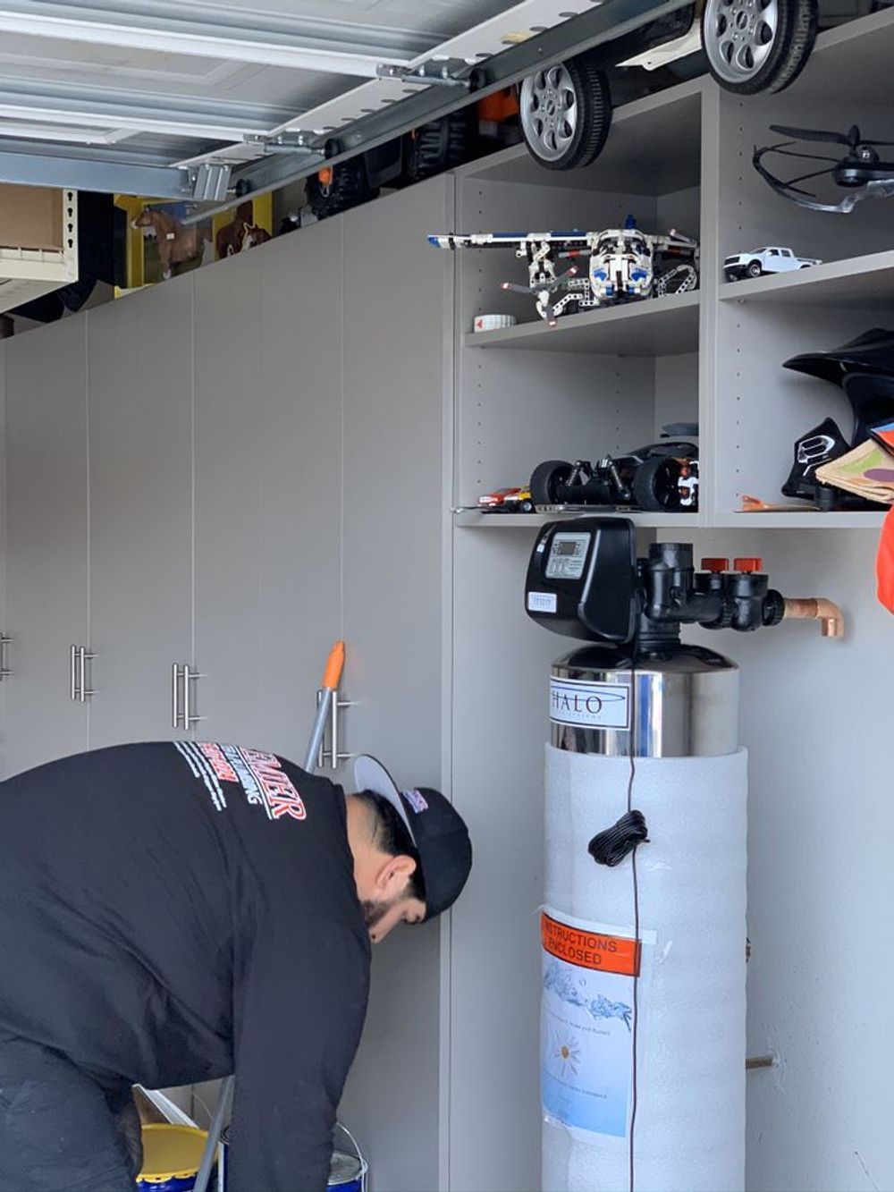Man examining water softener in a garage, cabinets and shelves with toys.