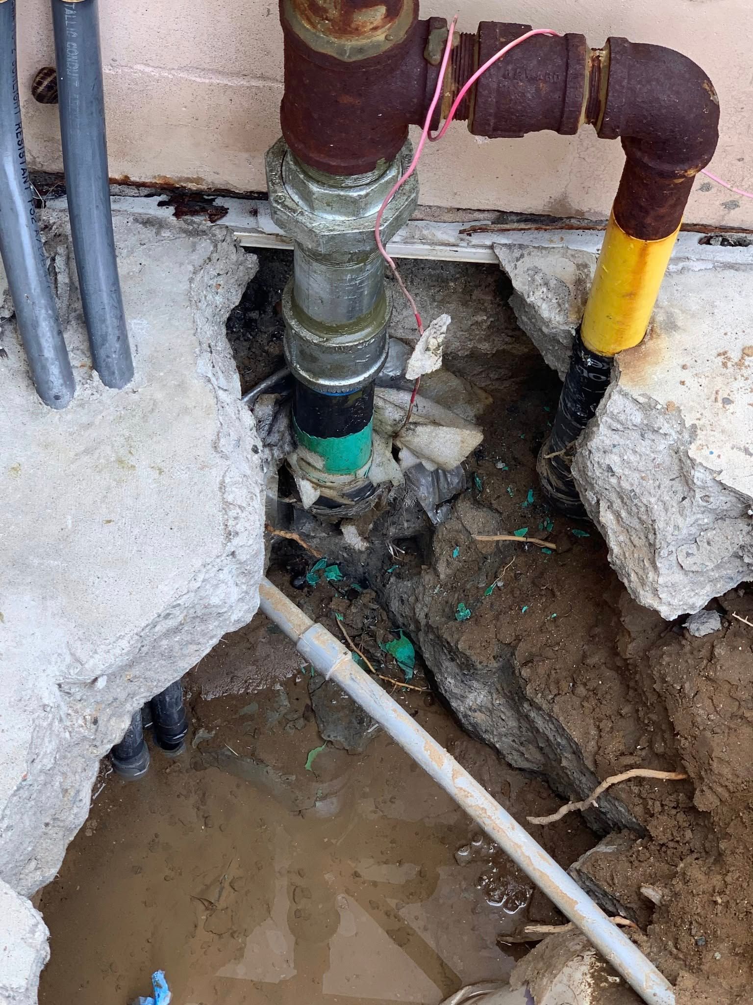 Pipes and fittings exposed in a hole in the concrete, showing a gas line, with various colors and dirt.