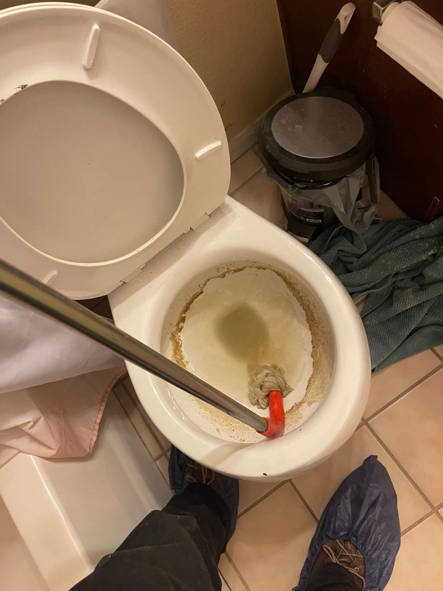 A toilet being unclogged with a snake tool, seen from above. A person wears blue shoe covers.