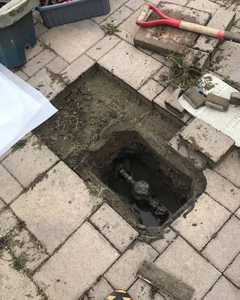 Open excavation in brick patio, revealing water meter and pipes, with tools nearby.