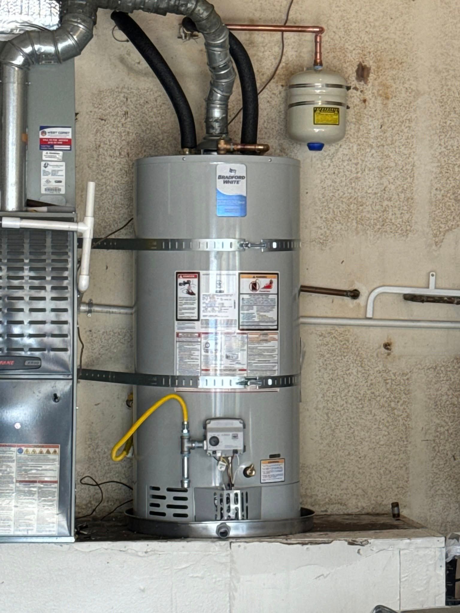 A gray water heater with a black pipe, copper piping, and an expansion tank in a utility room.