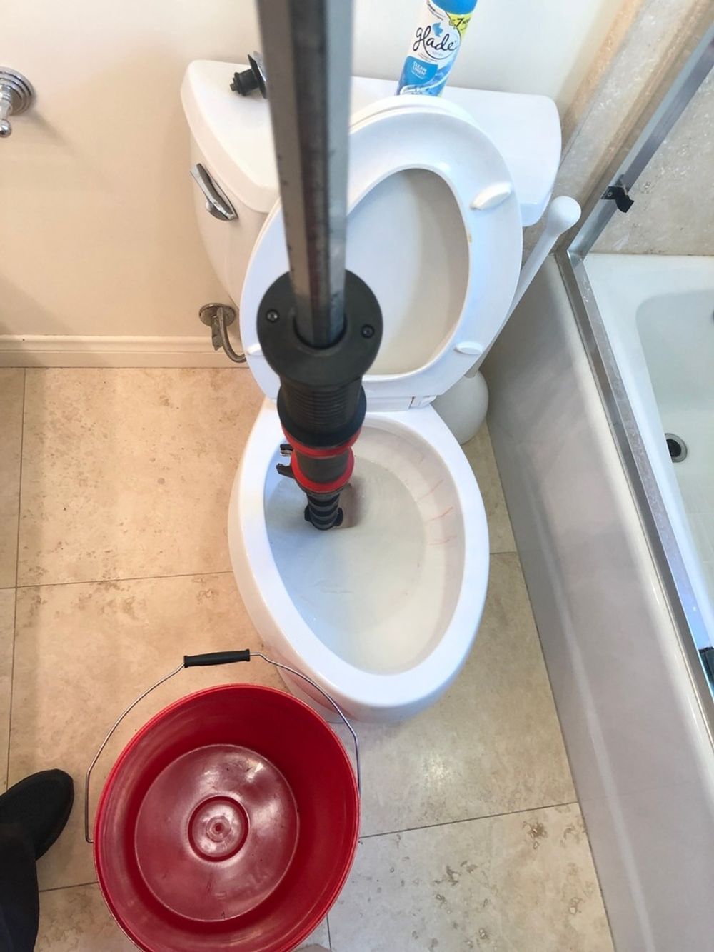 Person plunging a toilet in a bathroom with a red bucket. The toilet is white, and the floor is tiled.