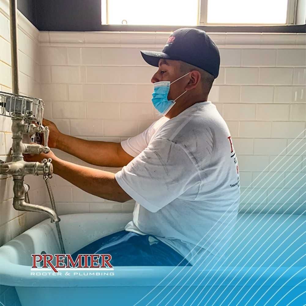 Plumber fixing tub faucet; wearing a mask and baseball cap, in a bathroom with white tiles.