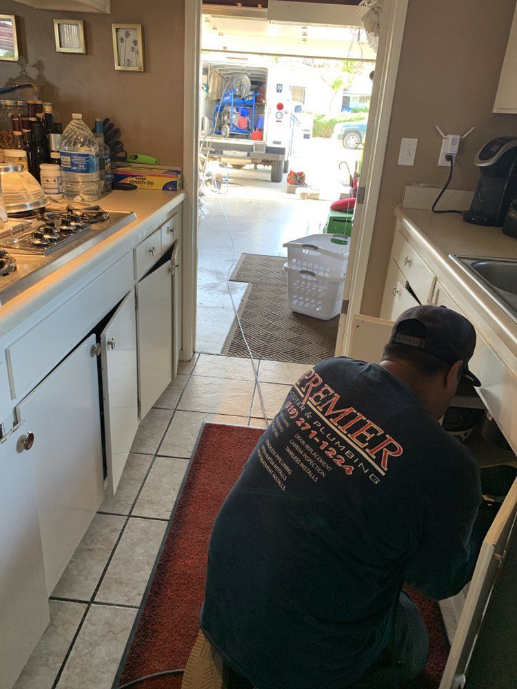 Man working on kitchen appliance, open door to outdoors. White cabinets, red rug, and tiled floor.