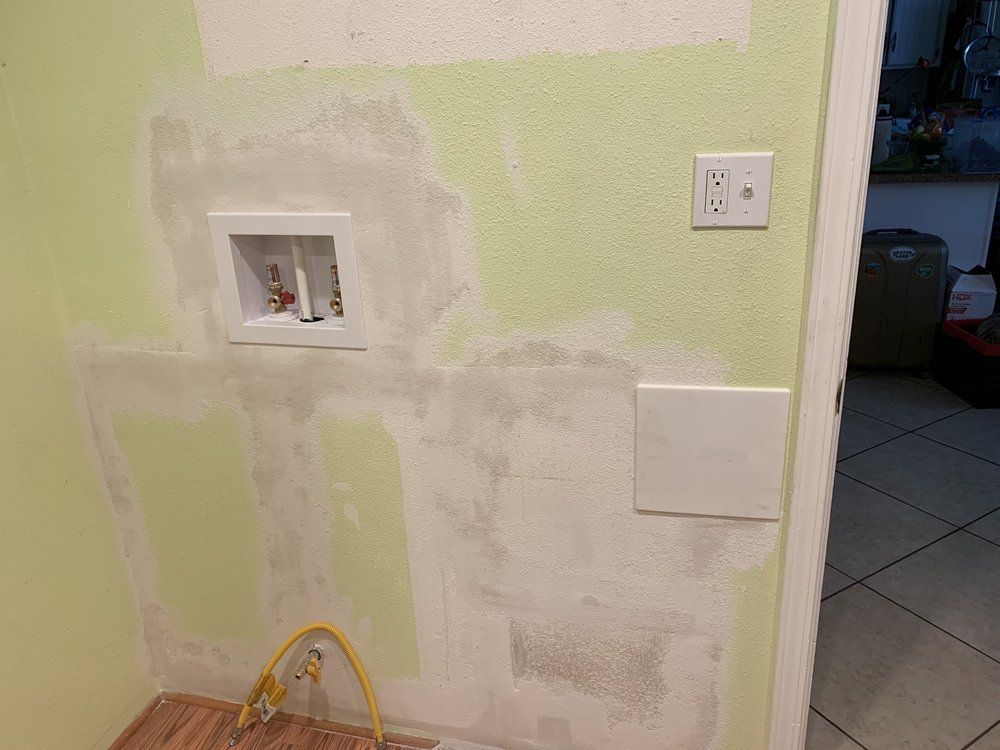 A wall with patched holes, an electrical outlet, laundry hookup box, and square access panel, ready for paint.