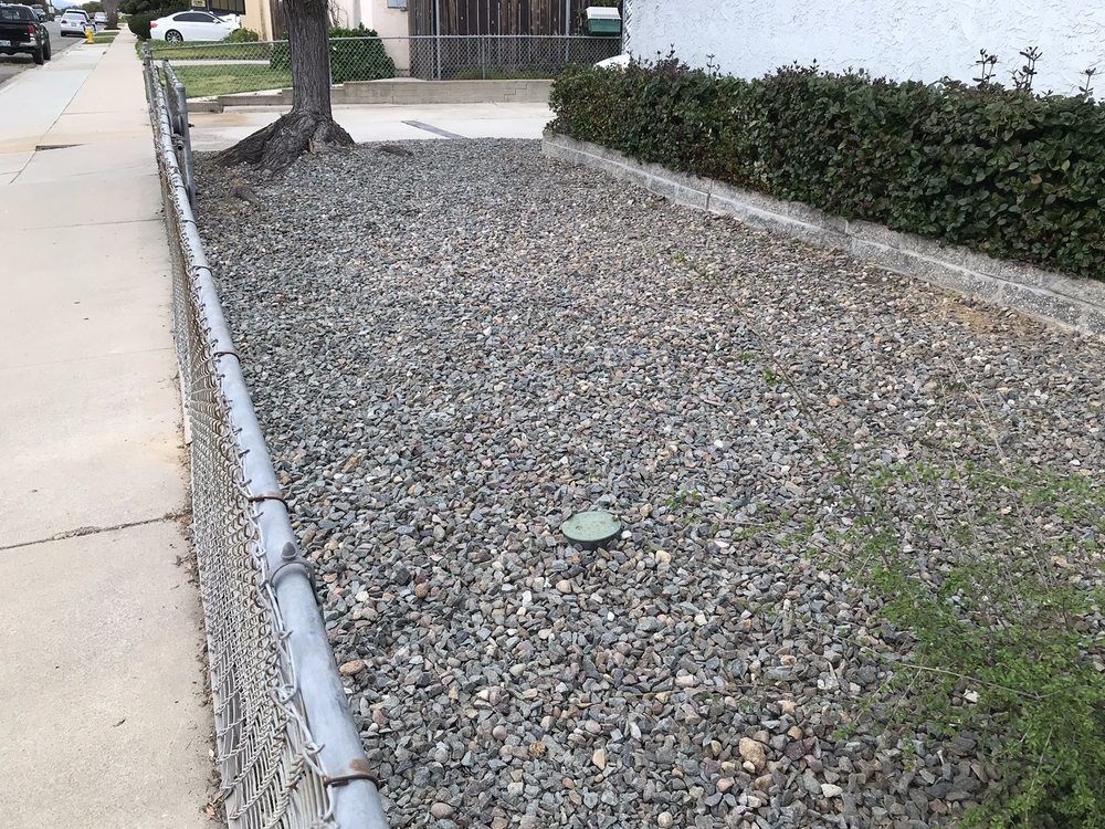 Gravel ground cover in a strip between a sidewalk and a chain-link fence, with green bushes along the back.