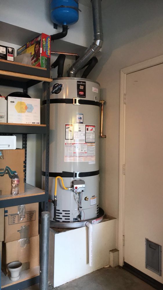 Water heater in a garage, next to shelves with boxes and a door. Copper and gray pipes visible.