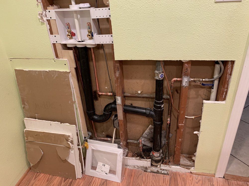 Plumbing exposed inside a wall. Black drain pipes, copper water pipes, and a white washing machine box are visible.