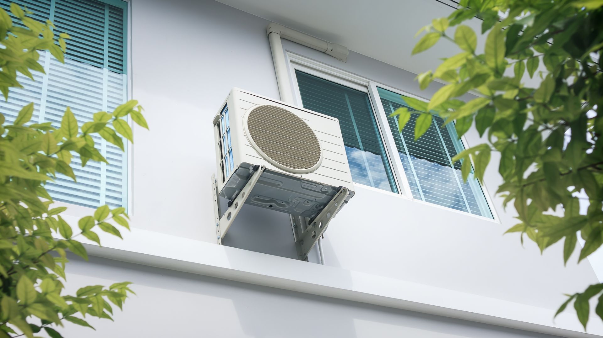 Outdoor air conditioner unit mounted beneath a window on a white building, partly framed by green leaves.
