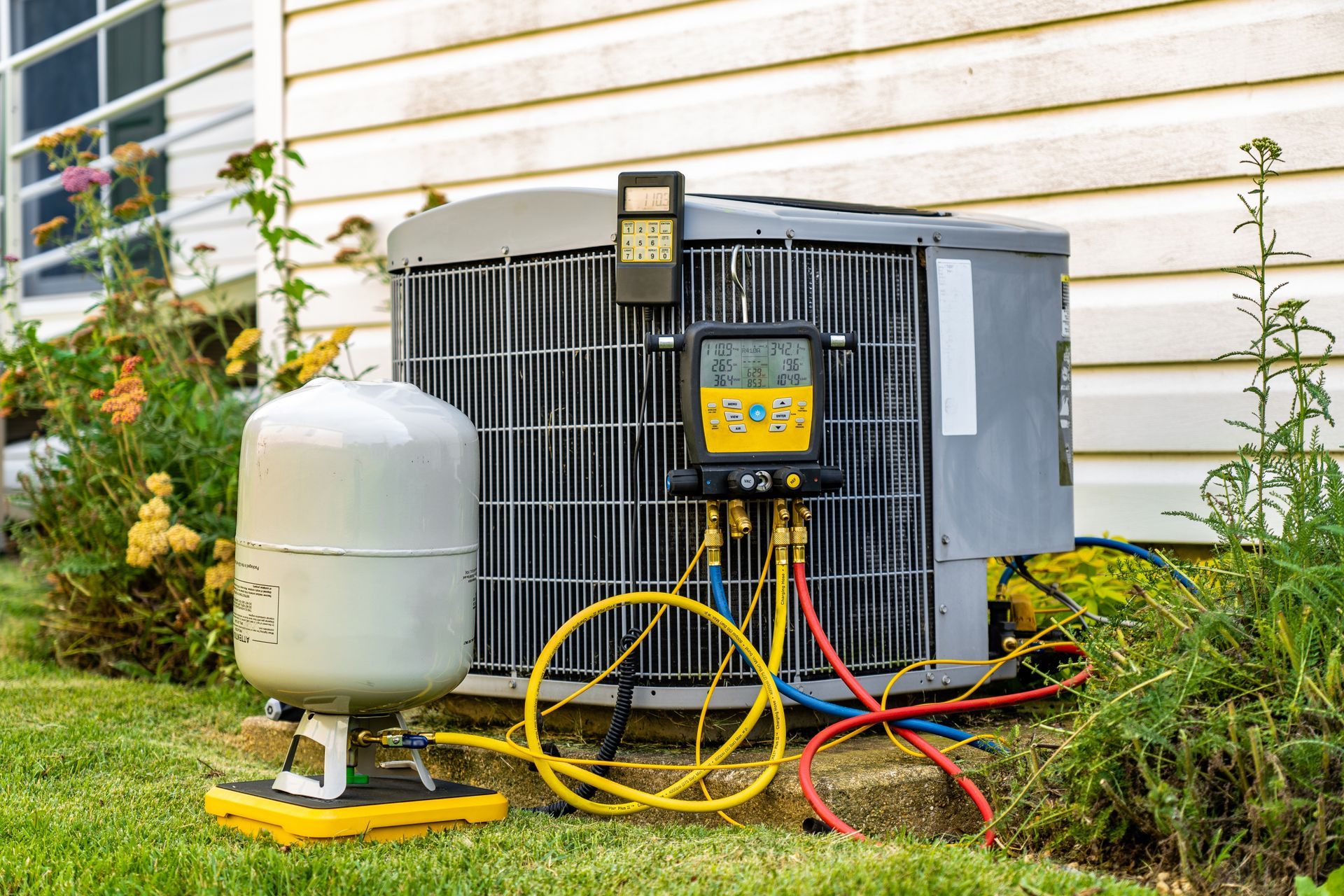 Outdoor HVAC condenser unit with gauges and hoses beside a house, next to a white tank on a grassy lawn