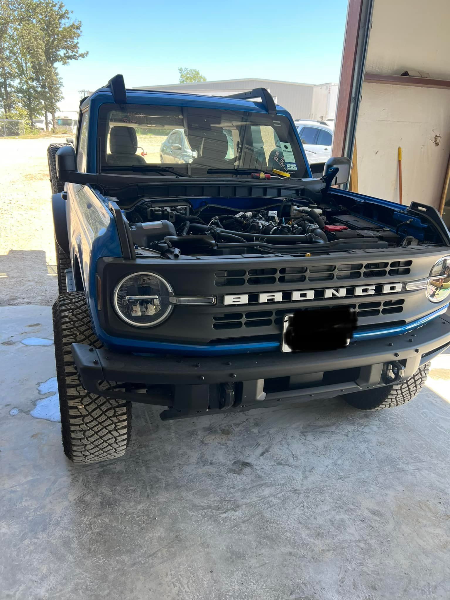 Blue Ford Bronco with hood open, parked outdoors in sunlight.