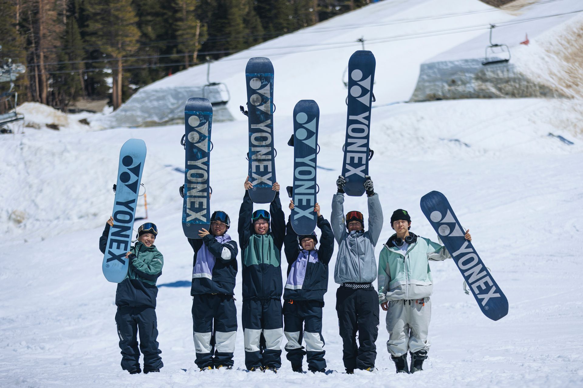Athleten halten Snowboards an einem verschneiten Berghang hoch. Auf den Boards ist „YONEX“ aufgedruckt.