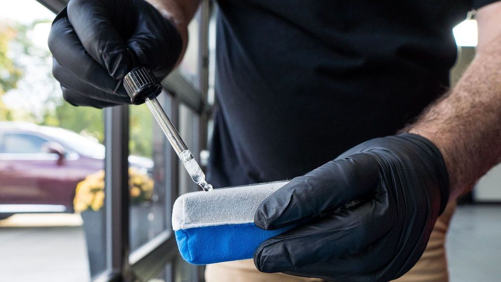 A person wearing black gloves applies a detailing product to a sponge applicator near a car.