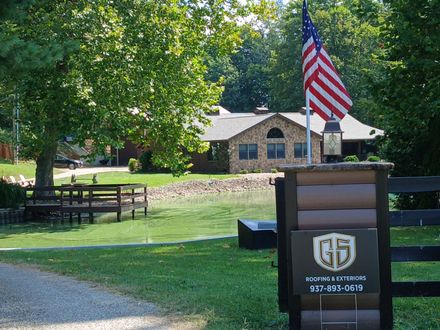 A brick house with a front deck sits behind a yard featuring a sign with the 