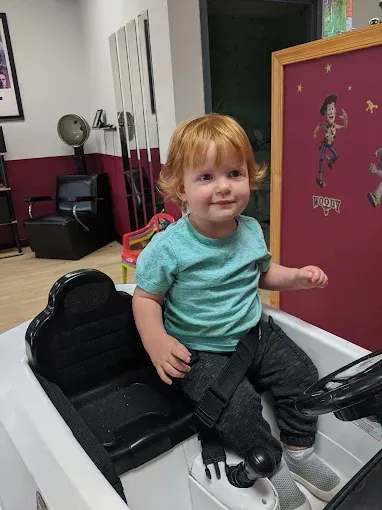 A little boy is sitting in a toy car at a barber shop.