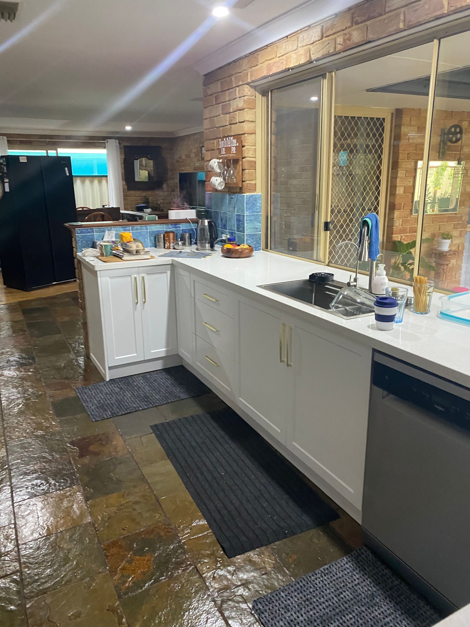 Kitchen with white cabinets, blue tile, and a brick wall, plus a window overlooking the yard.