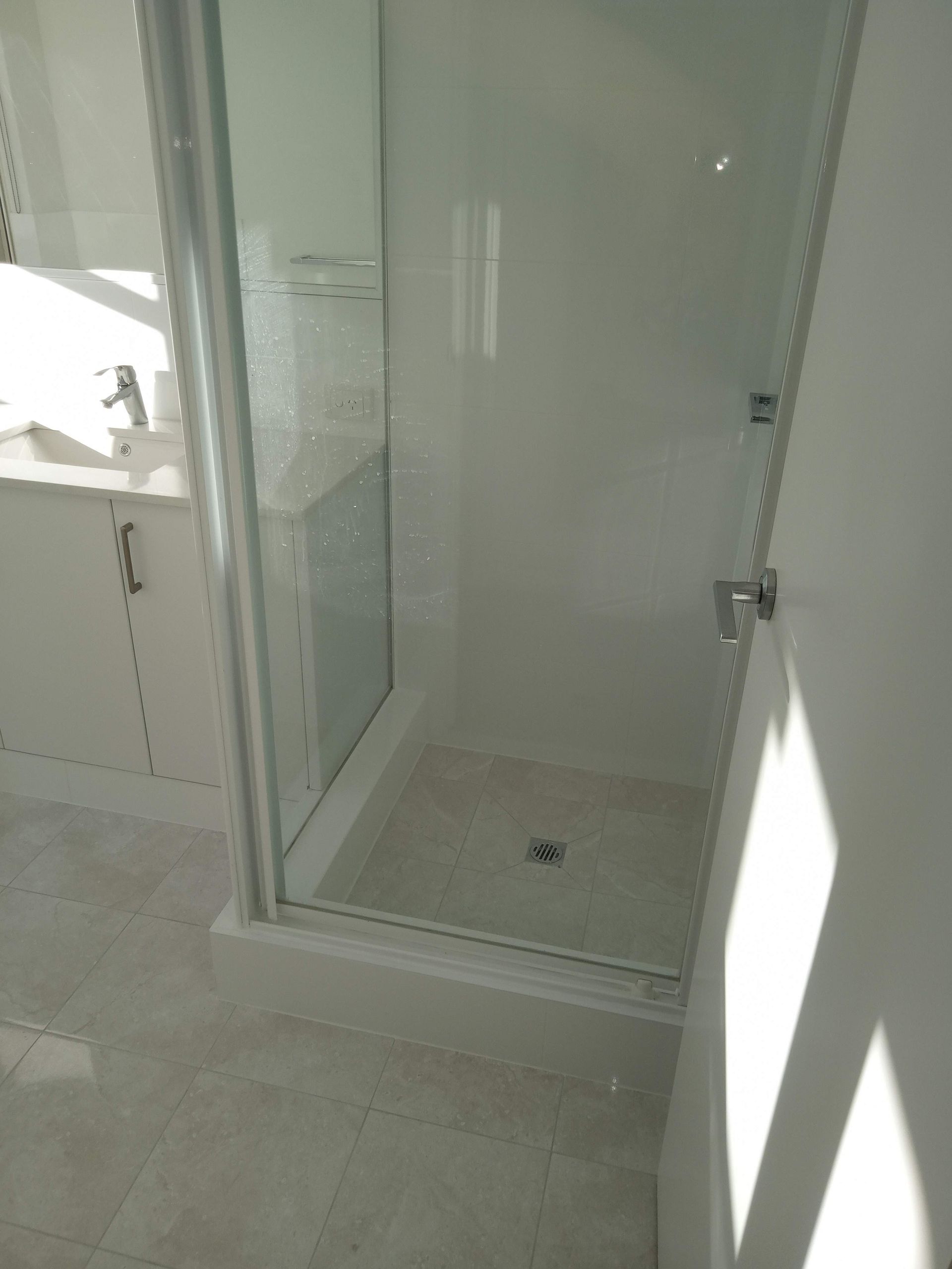 A small, white-walled shower with glass doors and a tiled floor; the sink is visible in the background.