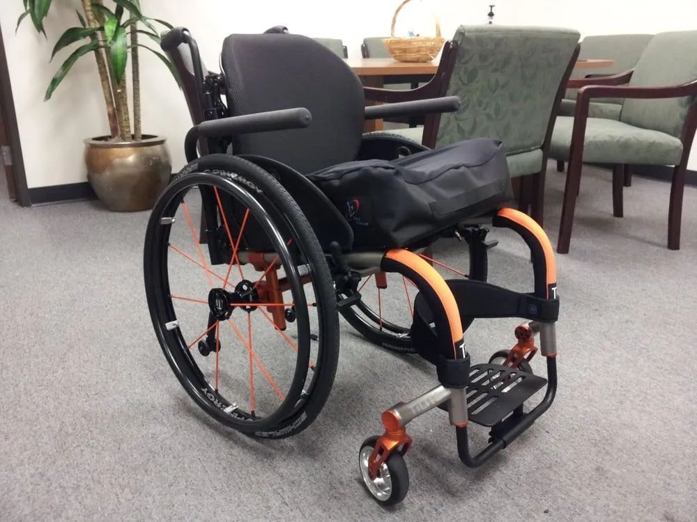 Modern wheelchair with black seat, orange accents, and black wheels, indoors.