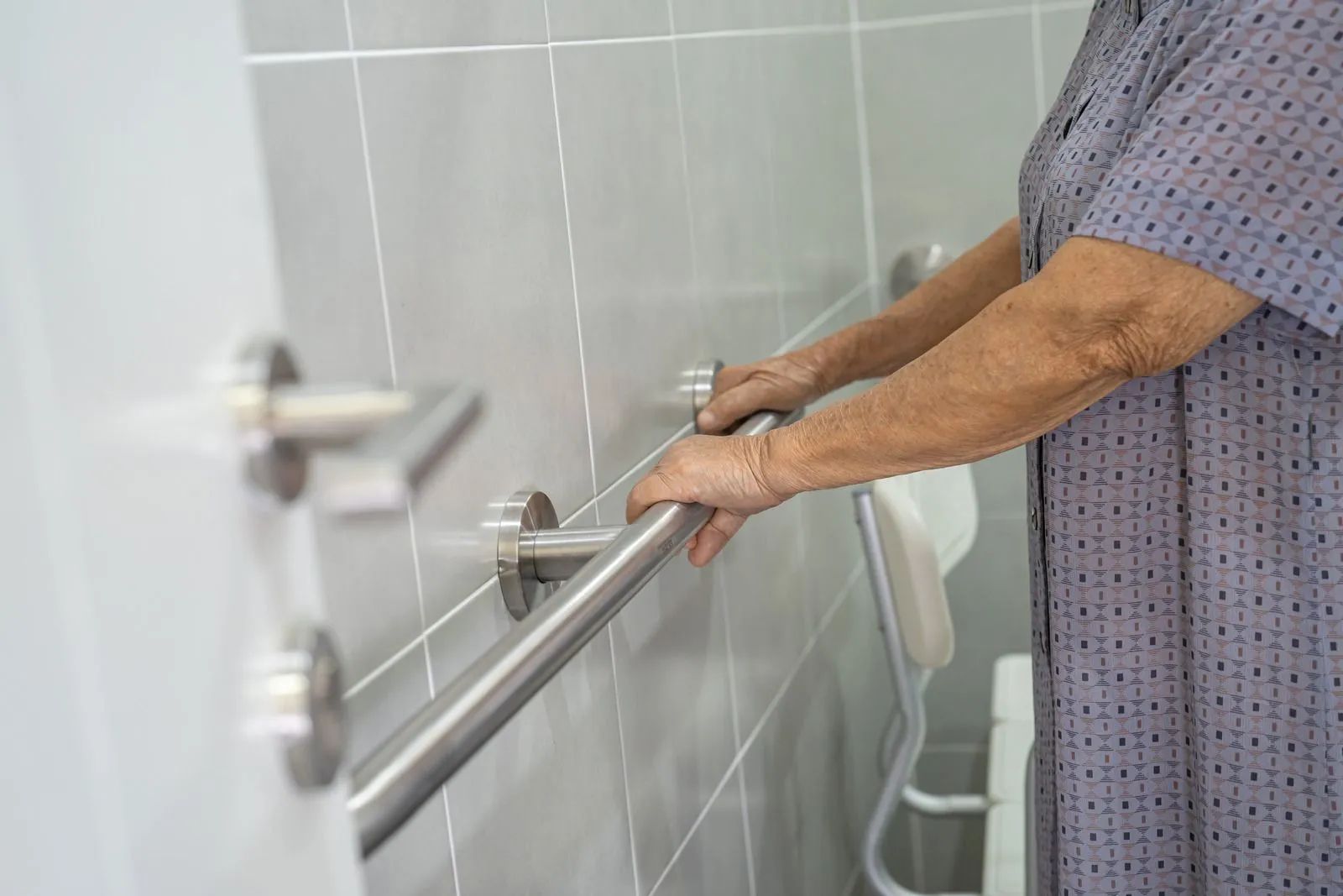Elderly person in a bathroom, using a handrail for support. Gray tiled wall, stainless steel
hardw