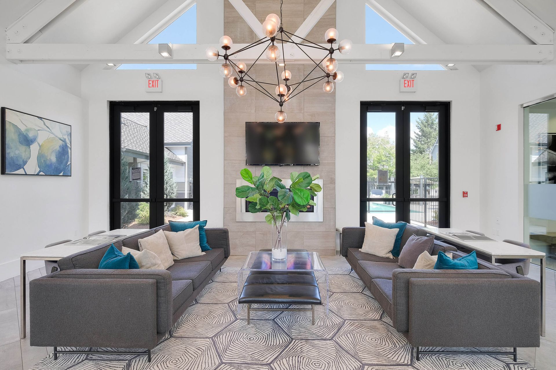 Bright communal lounge with two gray sofas, wall TV, chandelier, large windows, and a plant.