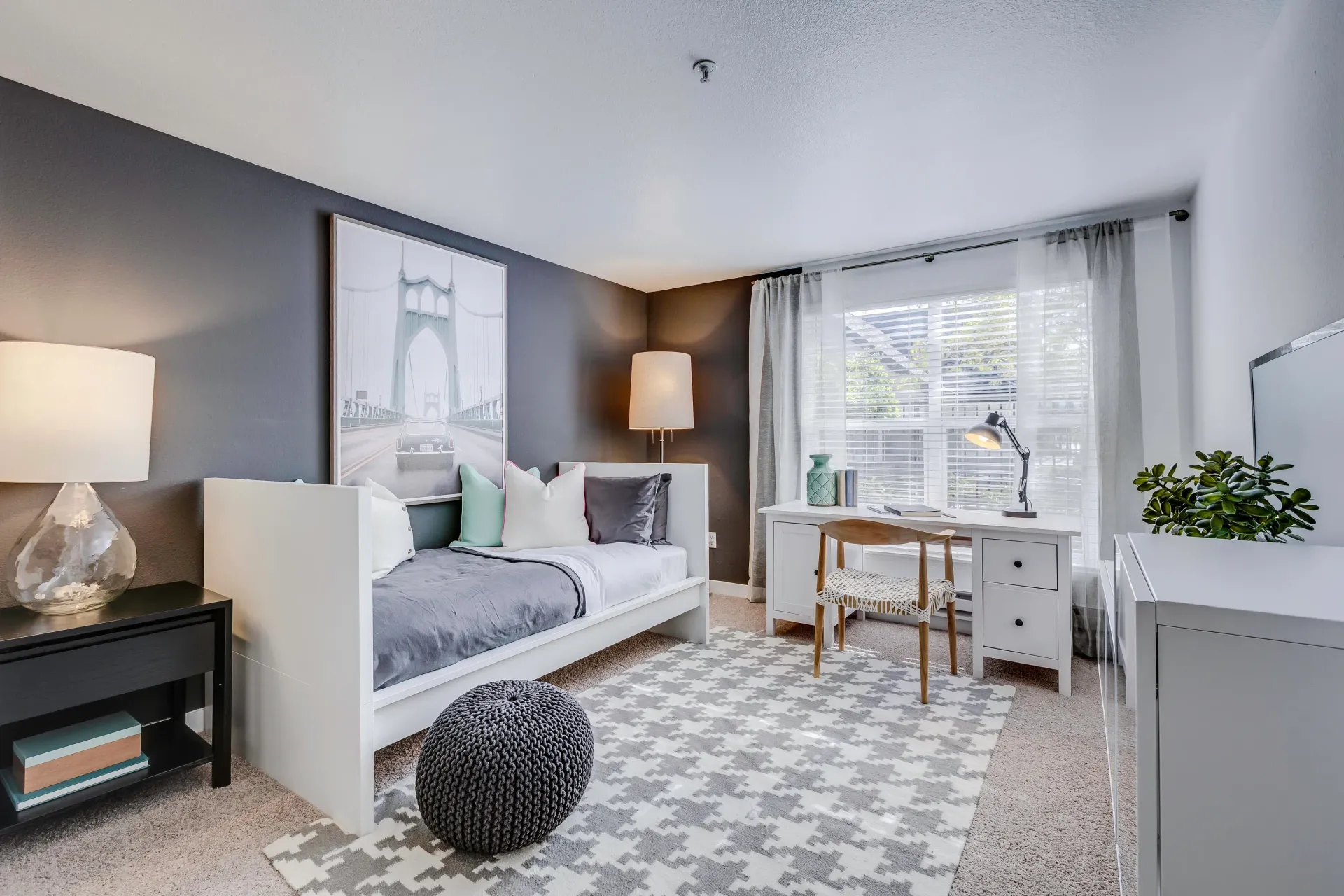 Bright modern bedroom in a white/gray apartment with a bed, desk, rug, and a window with sheer curtains.