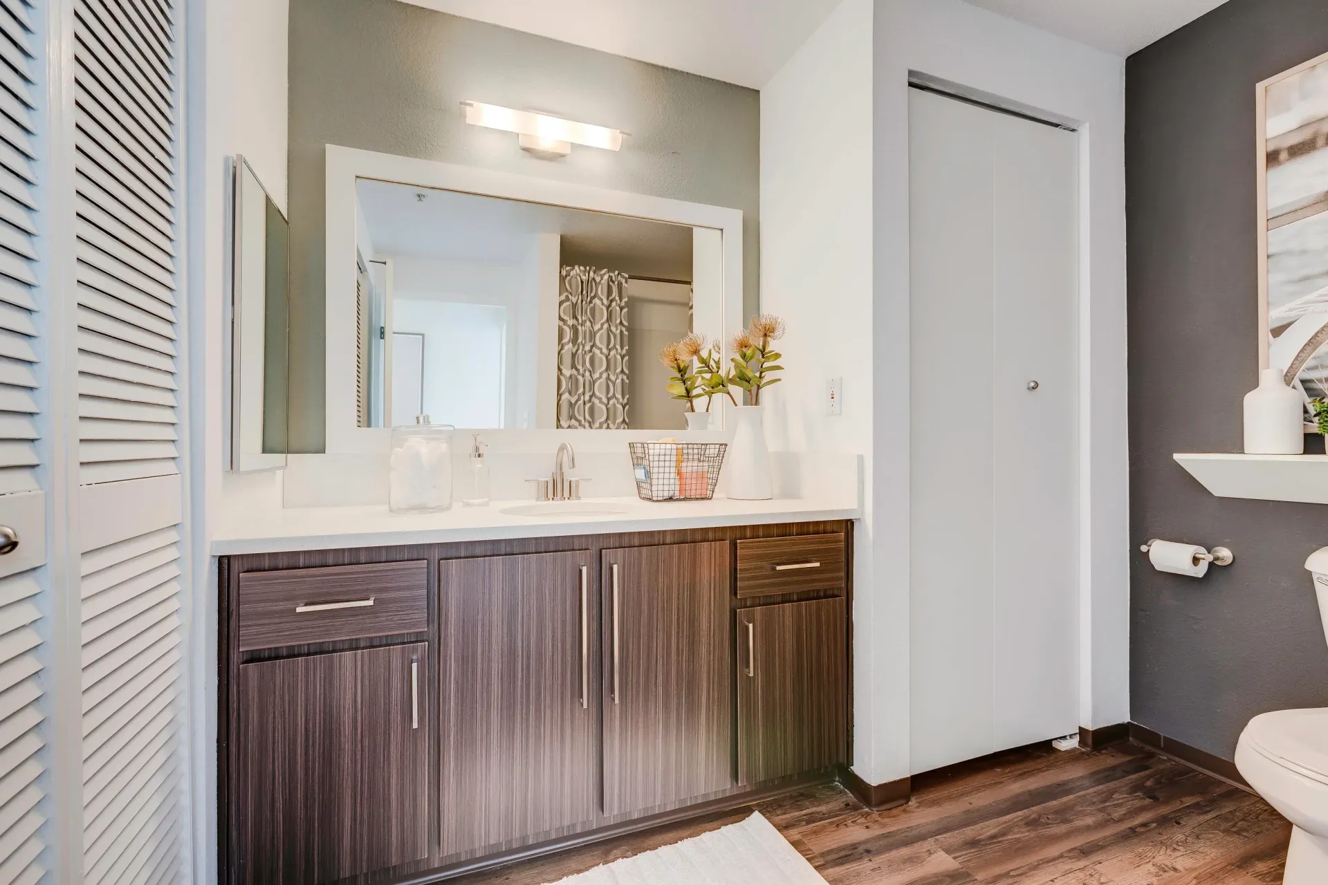 Bathroom vanity with white countertop, dark wood cabinets, large mirror, and toilet in a modern apartment.