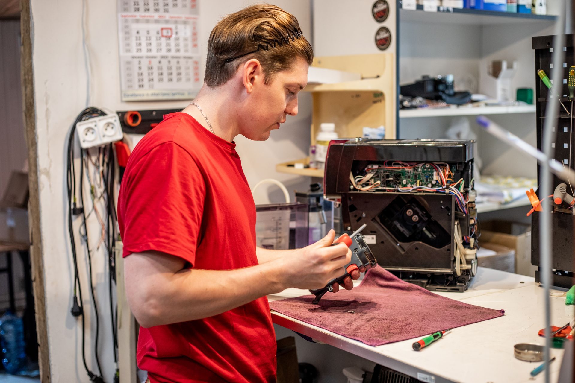 A man in a red shirt repairs electronics at a workbench. He's holding tools, focused on the open device.