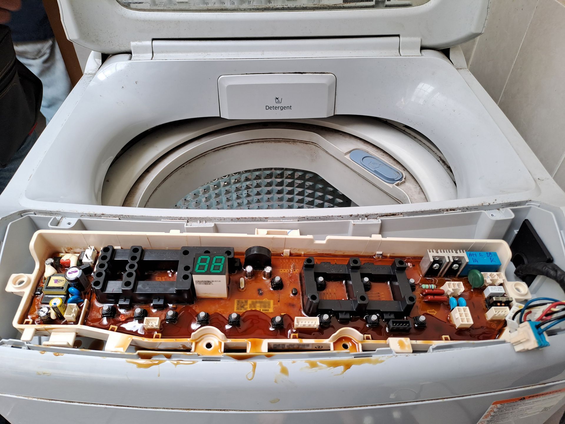 A top-load washing machine with its control panel exposed, revealing a circuit board with brown liquid residue.