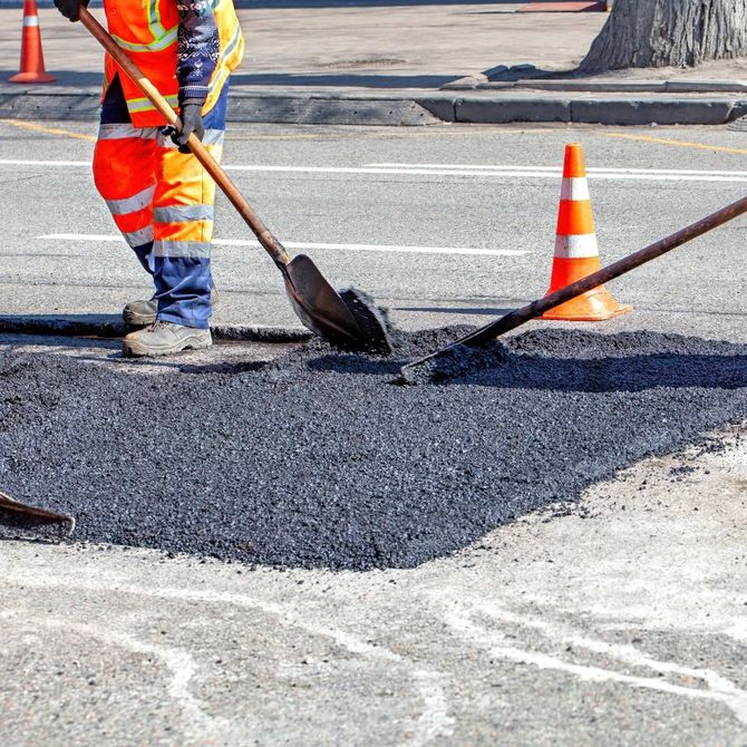 Road worker in high-visibility gear, shoveling asphalt on a street next to orange traffic cone.