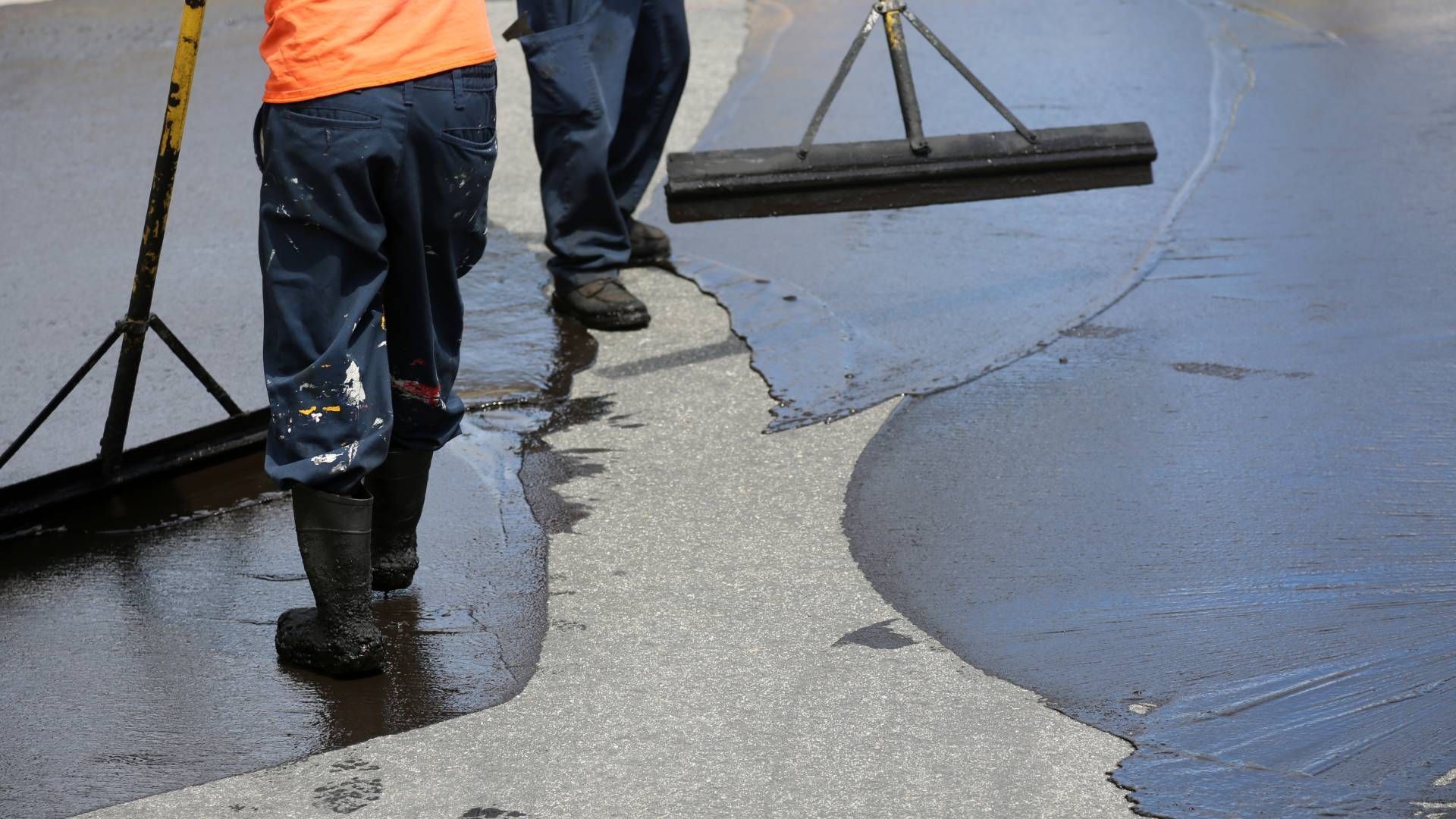 Two workers applying black sealant to a road with long-handled tools.