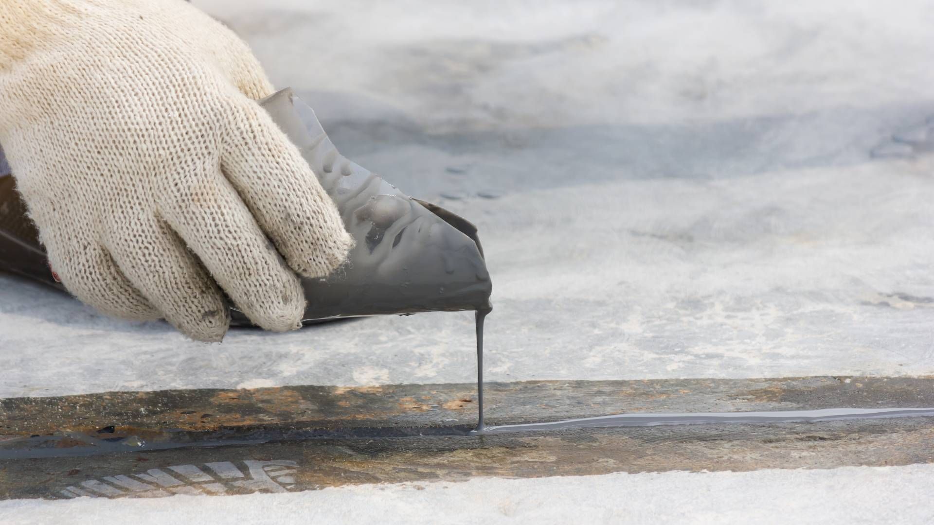 Gloved hand pouring black sealant into a seam on a light-colored surface.