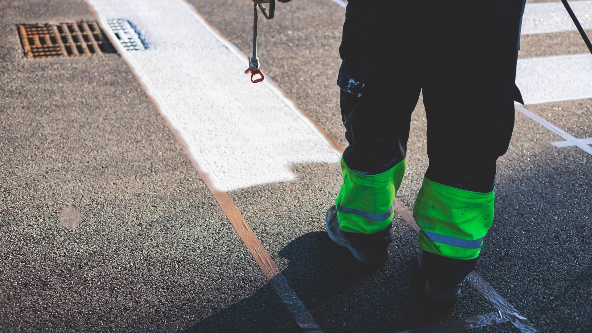 A worker standing on a road, painting white lines. They wear green high-visibility leg covers.
